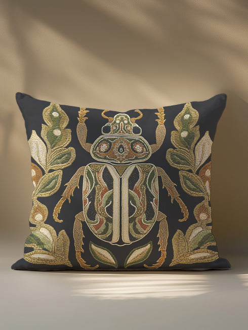 A square decorative throw pillow cover in a deep charcoal gray, almost black, fabric with a subtle linen texture. The pillow features a large, centrally-placed scarab beetle design rendered in a highly detailed, faux-embroidery style. The beetle is symmetrical, with intricate patterns in antique gold, olive green, muted orange, and cream covering its body and head. It is flanked by stylized botanical leaves in matching shades of green and gold.